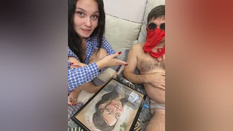 Video of a couple on a couch, woman with long black hair and blue shirt, man wearing red mask and glasses, holding framed photo.