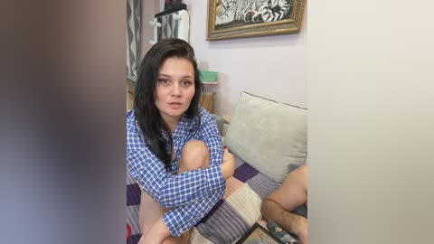 Video of a young woman with long dark hair, wearing a blue plaid shirt, sitting on a couch, holding a baby, in a cozy living room with a zebra print rug and framed artwork.