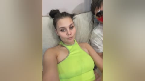 A video of a young woman with fair skin and dark hair styled in two buns, wearing a neon green halter top, lying on a white couch, partially obscured by a person in the background.