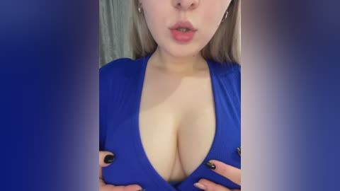 A close-up video of a light-skinned woman with long blonde hair, wearing a low-cut blue dress, lips parted, hands cupping her large breasts.