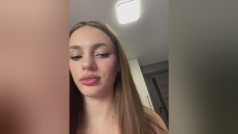 A video of a young Caucasian woman with long, straight brown hair and light skin, wearing makeup with pink lipstick, standing indoors near a ceiling light.