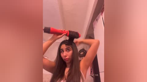 A young woman with long brown hair, wearing a white tank top, uses a red hair dryer in a bathroom with beige walls and a white shower curtain.