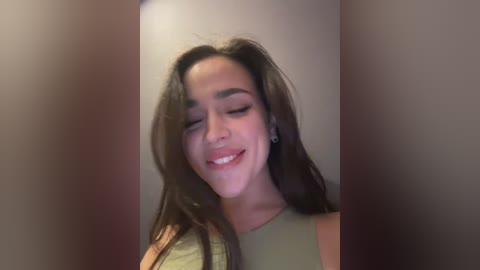 Video of a young woman with long, wavy brown hair, smiling and closing her eyes, wearing a sleeveless olive-green top, against a blurred, neutral-colored background.