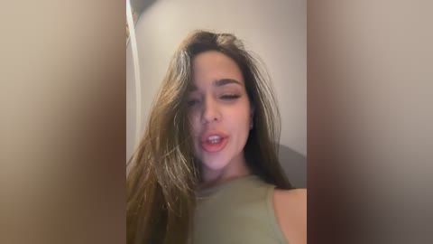 A video of a young woman with long, straight, dark brown hair, wearing a sleeveless green top, leaning forward with her eyes closed, lips parted, against a blurred beige background.