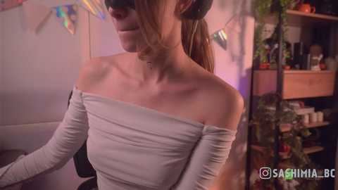 Video of a slender, fair-skinned woman with long brown hair in a ponytail, wearing a white, off-the-shoulder dress and a black mask, standing indoors in a cozy room with festive decorations.