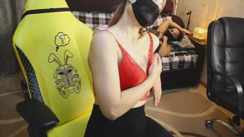 Video of a woman with fair skin and long brown hair, wearing a red lace bra and black skirt, sitting on a yellow gaming chair with bunny ears, holding a sleeping man in black clothes on a plaid sofa.