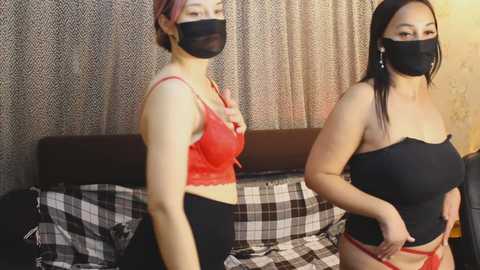 Video of two Asian women wearing black face masks, red lace bras, and black panties, standing in a bedroom with patterned curtains and a plaid bedspread.