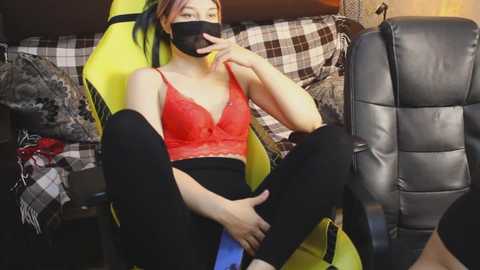 Video of a young Asian woman with fair skin, wearing a red lace bra, black leggings, and a black face mask, seated in a yellow gaming chair, surrounded by checkered-patterned cushions and a black leather couch.