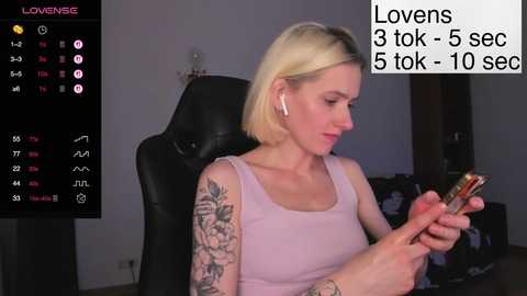 A video of a blonde woman with a pink tank top, tattoos, and white earbuds, sitting in a black chair, checking her phone.