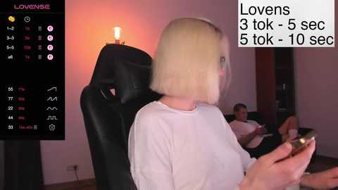 Video of a woman with a short blonde bob, wearing a white shirt, watching a TV screen displaying a man in a black chair. Background shows a dimly lit living room with a fireplace and another man sitting.