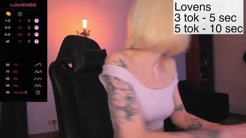 A blurry video of a blonde woman with tattoos, wearing a white tank top, sitting at a black chair in a dimly lit room. Text overlays include \"Lovens\" and \"3 tok-5 sec.\