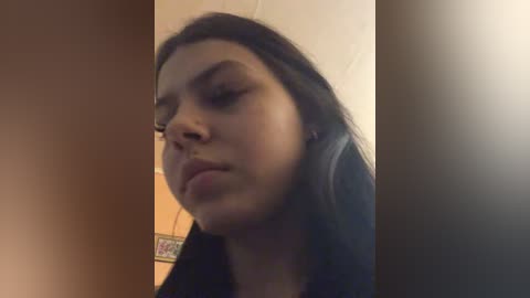 Video of a young woman with medium skin tone and long, dark hair, wearing a black top, and large black headphones. The background is a beige wall with a small decorative sticker.