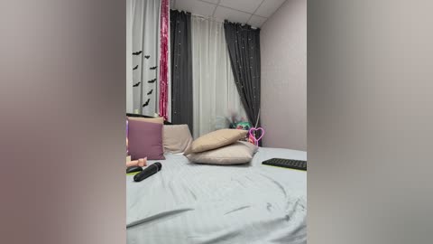 Video of a neatly made bed with light gray sheets, beige pillows, and a black remote control. Pink and white curtains cover the window.