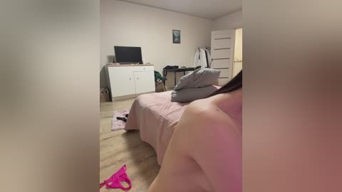A video of a dimly lit, beige room with a TV, desk, and bed, featuring a partially nude woman with fair skin, lying on a pink bedspread, wearing pink panties.
