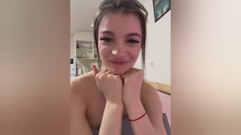 Video of a young, smiling woman with light skin and brown hair, topless, resting her chin on her hands, in a dimly-lit room with a white wall and a framed picture.