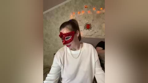Video of a woman with a red masquerade mask, white top, and pearls, seated indoors, with a soft, muted wallpaper background.