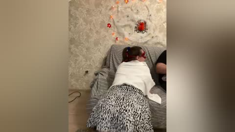 A video captures a young child, lying on a massage table with a white top and black and white patterned skirt, while an adult, partially visible, performs a massage. The room has a cream-colored wall with a floral pattern and warm lighting.