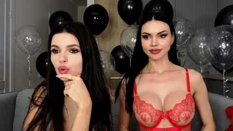Video of two women with long black hair, fair skin, and large breasts; one wears a sheer red lace bra, the other is topless. Background includes black and silver balloons, a wooden wall, and a gray couch.