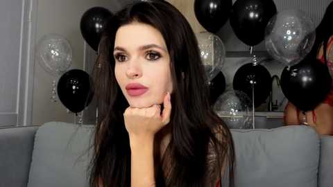 Video of a fair-skinned woman with long, straight black hair, wearing red lipstick, resting her chin on her hand, surrounded by black and silver balloons in a modern, brightly lit room.
