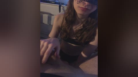 A dimly lit video of a young woman with long brown hair, wearing a black bra and panties, sitting on a bed. She has fair skin and is blindfolded.