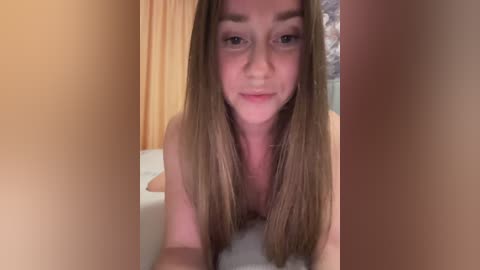 A video of a nude young woman with fair skin and long brown hair, lying on a bed in a dimly lit room.