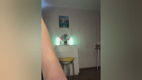 A video of a dimly-lit, sparsely decorated room with a white fireplace, a yellow stool, and a blue painting on the wall.