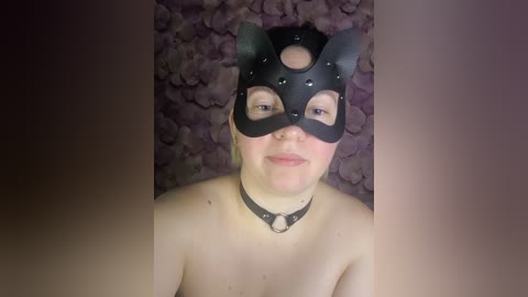 Video of a fair-skinned, topless woman with a black leather cat mask and collar, against a floral patterned background.