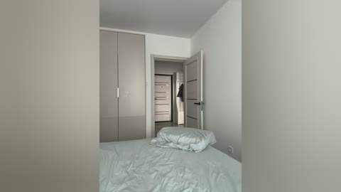 A video of a minimalist, modern bedroom with light grey walls and furniture. The bed is made with white sheets, and a person in black pants and a white shirt is standing in an adjacent doorway.