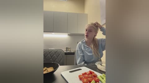 Video of a blonde woman with fair skin, wearing a blue hoodie, sitting at a modern kitchen table with tomato slices and crackers.