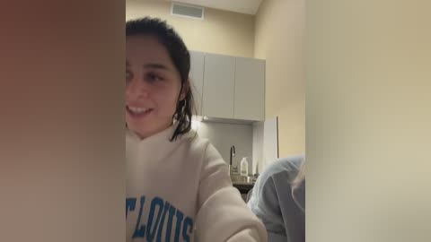 Video of a smiling young woman with dark hair, wearing a white \"CLOTHES\" sweatshirt, in a dimly lit, beige-walled room with a kitchenette in the background.