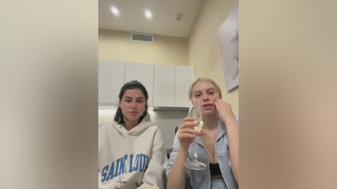 A video of two young women in a beige-walled room, both wearing hoodies. One holds a drink, the other rests her head on her hand.