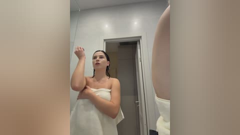 Video of a fair-skinned woman with wet hair, wrapped in a white towel, standing in a modern, minimalist bathroom with a large mirror reflecting her body.