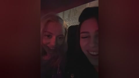 Video of two women smiling, one with blonde hair, the other with dark hair, both in black clothing. Background features a dimly lit, tiled wall.