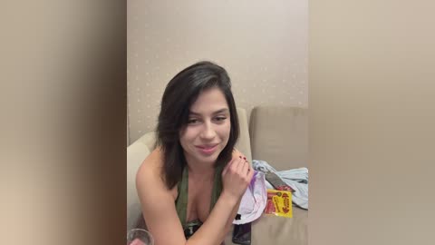Video of a smiling young woman with medium-length dark hair, wearing a green halter top, sitting on a beige couch, holding a yellow envelope, with a light-colored wall in the background.