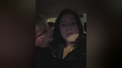 A dimly lit video of a young woman with shoulder-length black hair, wearing a black leather jacket, sitting in a dimly lit car, holding a phone to her ear.