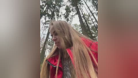 A blurry video of a young woman with long blonde hair, wearing a bright red jacket, standing amidst tall, dense trees in a forest.