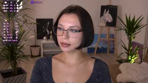 Video of a young, fair-skinned woman with short black hair and glasses, wearing a gray top, standing in an artist's studio with plants, paintings, and a laptop.