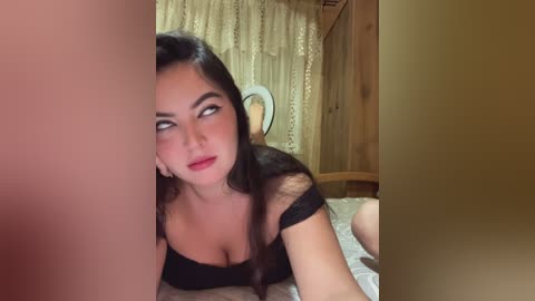 Video of a young woman with long dark hair and fair skin, wearing a black lace top, lying on a bed with white sheets, looking to the left. The background features wooden walls and a white curtain.