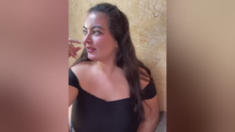 Video of a young woman with long dark hair, wearing a black off-shoulder top, standing against a beige textured wall, with a blurred, pink background.