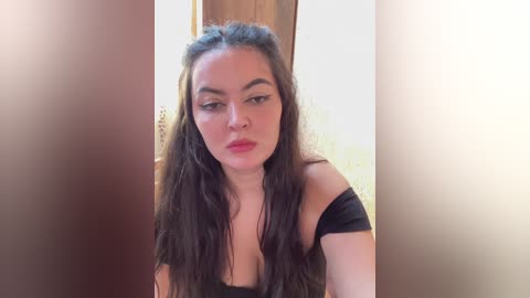 Video of a young woman with long, wavy dark hair and fair skin, wearing a black off-shoulder top, looking introspective, set against a blurred wooden background.