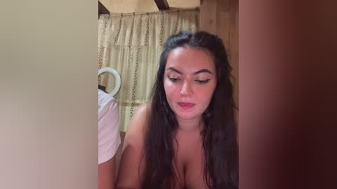 Video of a young woman with long, dark hair and light skin, wearing minimal makeup, leaning against a doorway. The background shows a wooden interior with a beige curtain and a white object.