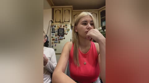 Video of a young blonde woman in a bright pink tank top, pensive, sitting in a cluttered kitchen with a refrigerator door covered in photos and magnets.