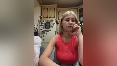 A video of a young blonde woman in a sleeveless red top, sitting pensively, hand on chin, in a cozy, cluttered kitchen with wooden cabinets and a refrigerator covered in magnets.