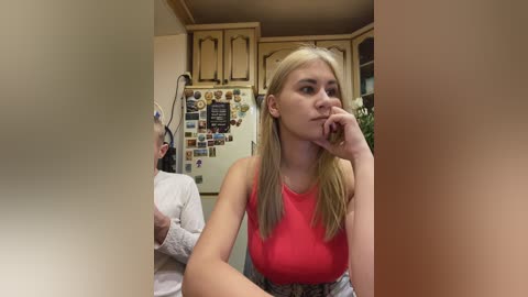Video of a blonde young woman in a red tank top, sitting pensively in a rustic kitchen, surrounded by wooden cabinets and a refrigerator covered in colorful magnets.