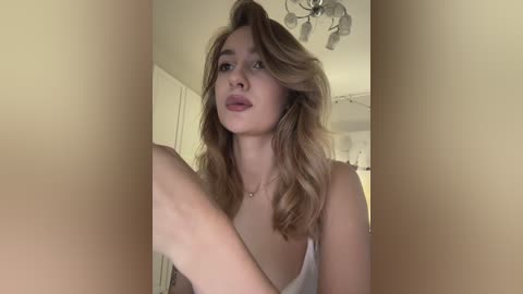 Video of a young Caucasian woman with long, wavy blonde hair, wearing a white tank top, standing indoors with a soft, beige background.