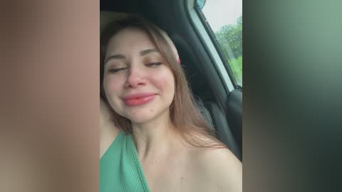 Video of a young Caucasian woman with light skin and long brown hair, wearing a green tank top, sleeping with eyes closed in a car seat.