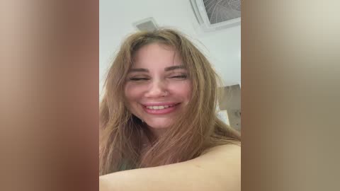 Video of a smiling, fair-skinned woman with long, light brown hair, wearing a nose ring, and topless, partially obscured by two brown objects, possibly hands, in a bright, indoor setting with white walls and a ceiling fan.