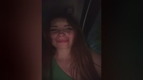 A dimly lit video shows a young woman with long, wavy brown hair, smiling. She wears a green tank top, and the background is dark and indistinct.