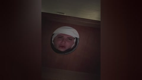Video of a man's head with a white bandage wrapped around it, peering through a circular hole in a wooden door, dimly lit, suggesting a suspenseful or hidden setting.