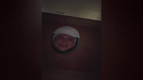 A dimly lit video showing a smiling face inside a wooden door, partially obscured by a white helmet, with the door's grain visible in the background.